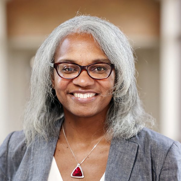 Headshot of a person with shoulder-length gray hair wearing a gray blazer, cream top, and a large triangular red pendant necklace, posed indoors with a softly blurred background.