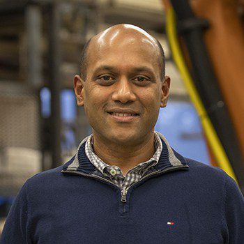 Headshot of a person with a shaved head wearing a navy blue zip-up pullover over a checked collared shirt, posed in an industrial setting with machinery and equipment in the background.