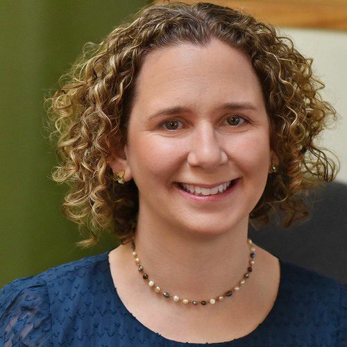 Headshot of a person with short, curly light brown hair, wearing a blue textured top and a beaded necklace, seated indoors with a light green background.