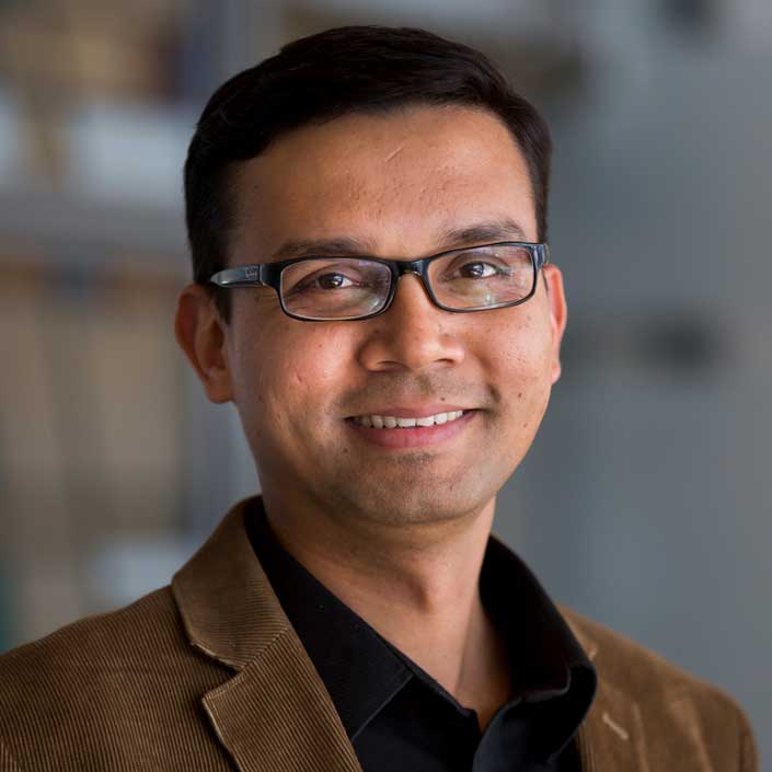 Headshot of a person with short dark hair wearing a brown corduroy blazer over a black collared shirt, posed indoors with a softly blurred background.