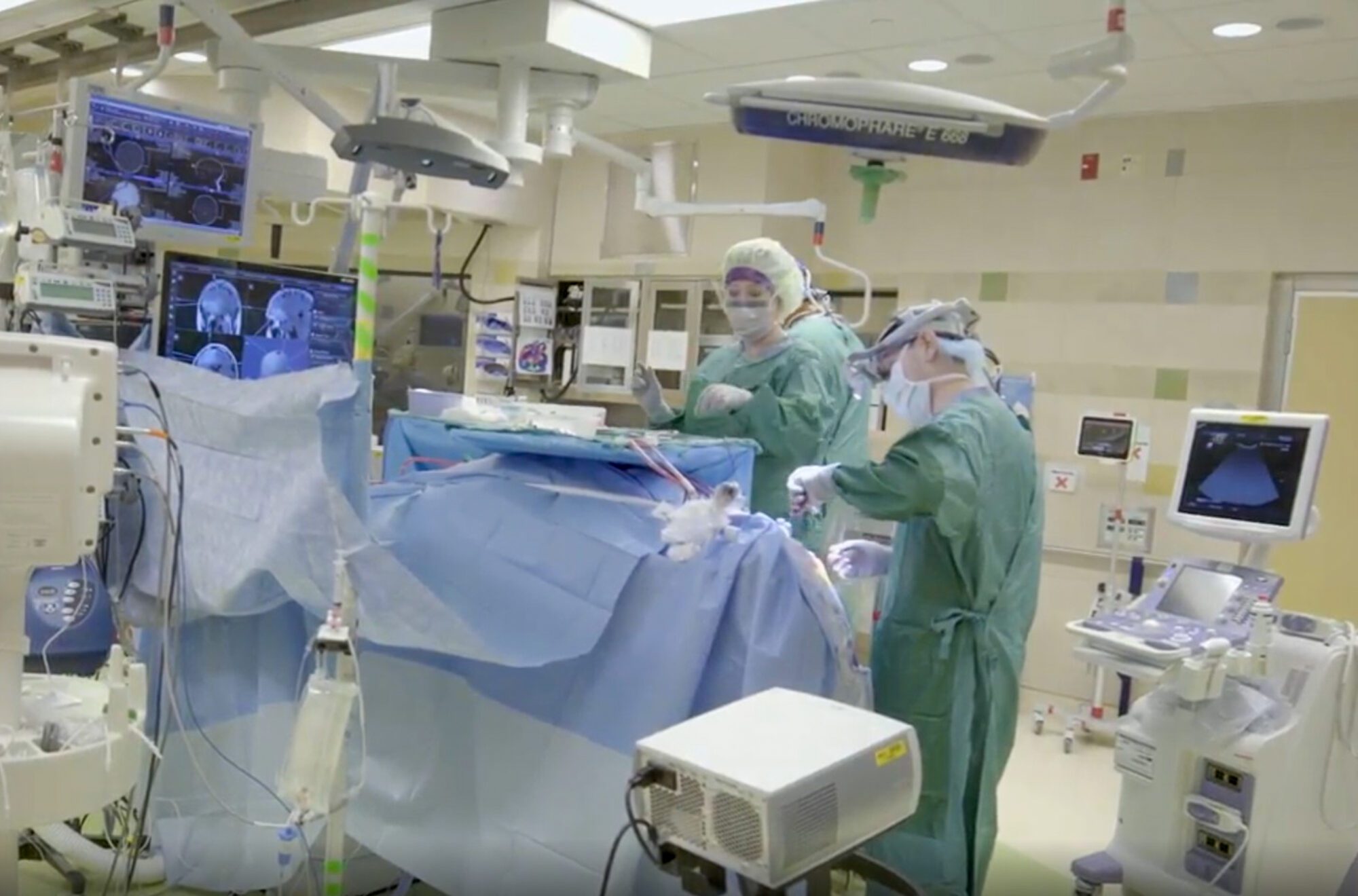 A surgical team in green scrubs performs a procedure in an operating room surrounded by medical equipment and monitors displaying brain scans.