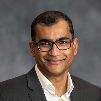 Person wearing a dark suit jacket over a white collared shirt, posed in front of a gray, softly blurred background.