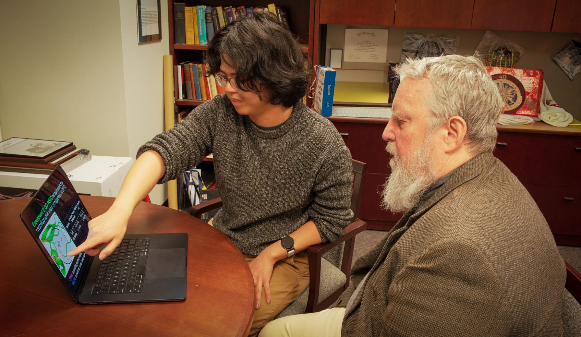 Two people sit at a table in an office; one is pointing at a laptop screen displaying slides with scientific diagrams. Books and awards are visible on shelves in the background.