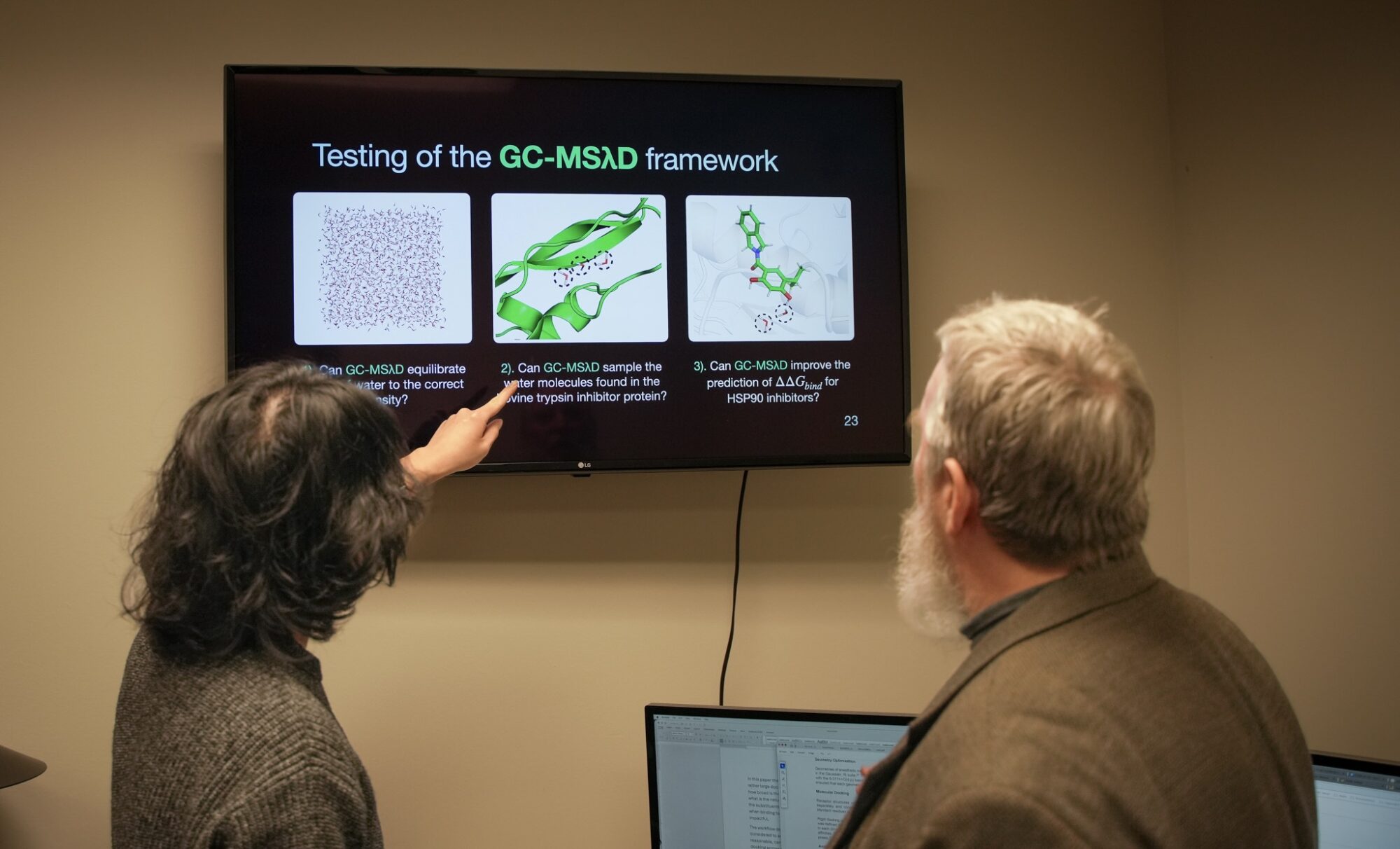 Two people are standing in an office, looking at a wall-mounted screen displaying scientific slides titled "Testing of the GC-MSAD framework," featuring molecular and protein graphics. One person is pointing at the screen.