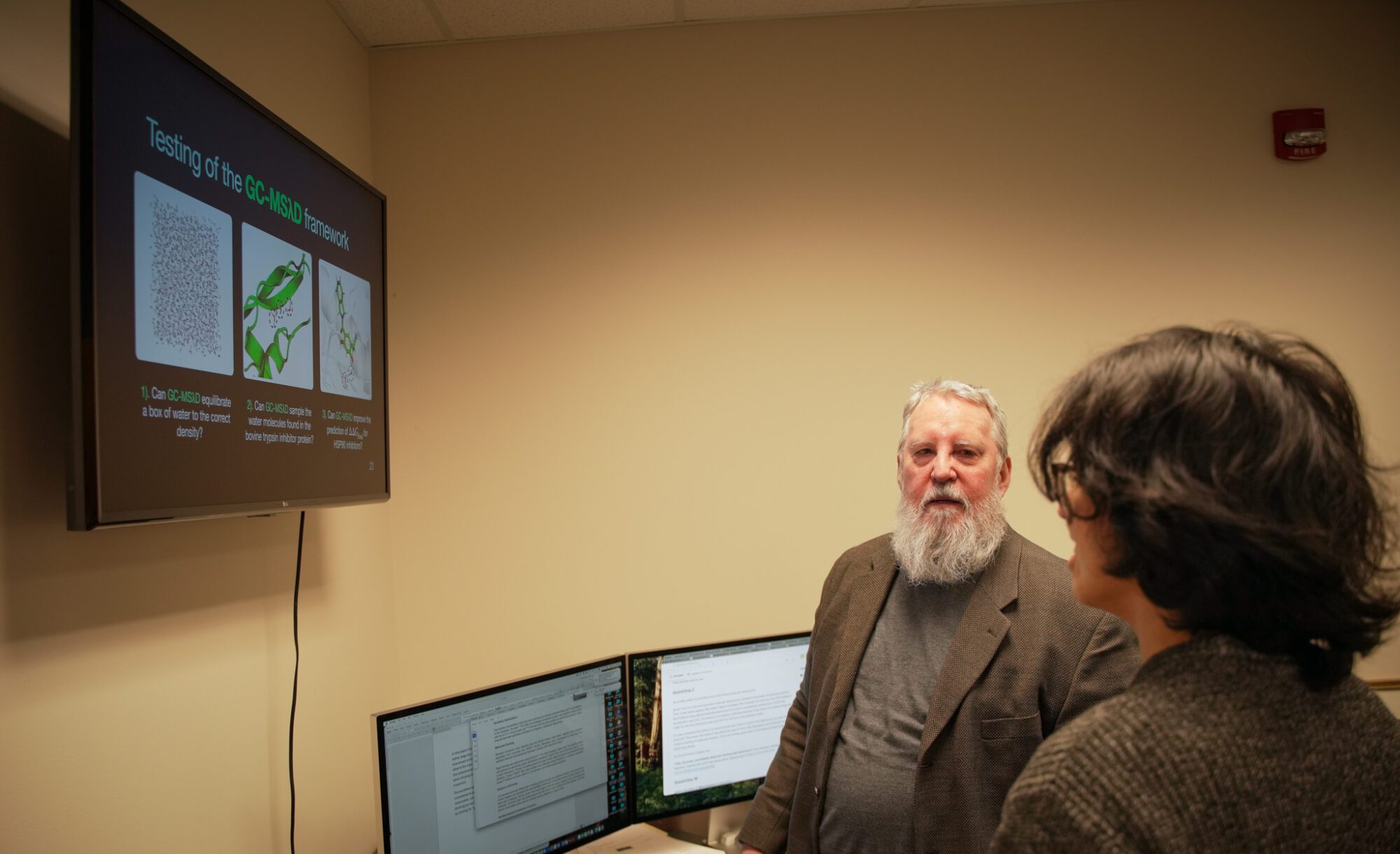 Two people in an office view a wall-mounted screen showing science slides titled "Testing of the GC-MSAD framework." Multiple computer monitors are on the desk beneath the screen.