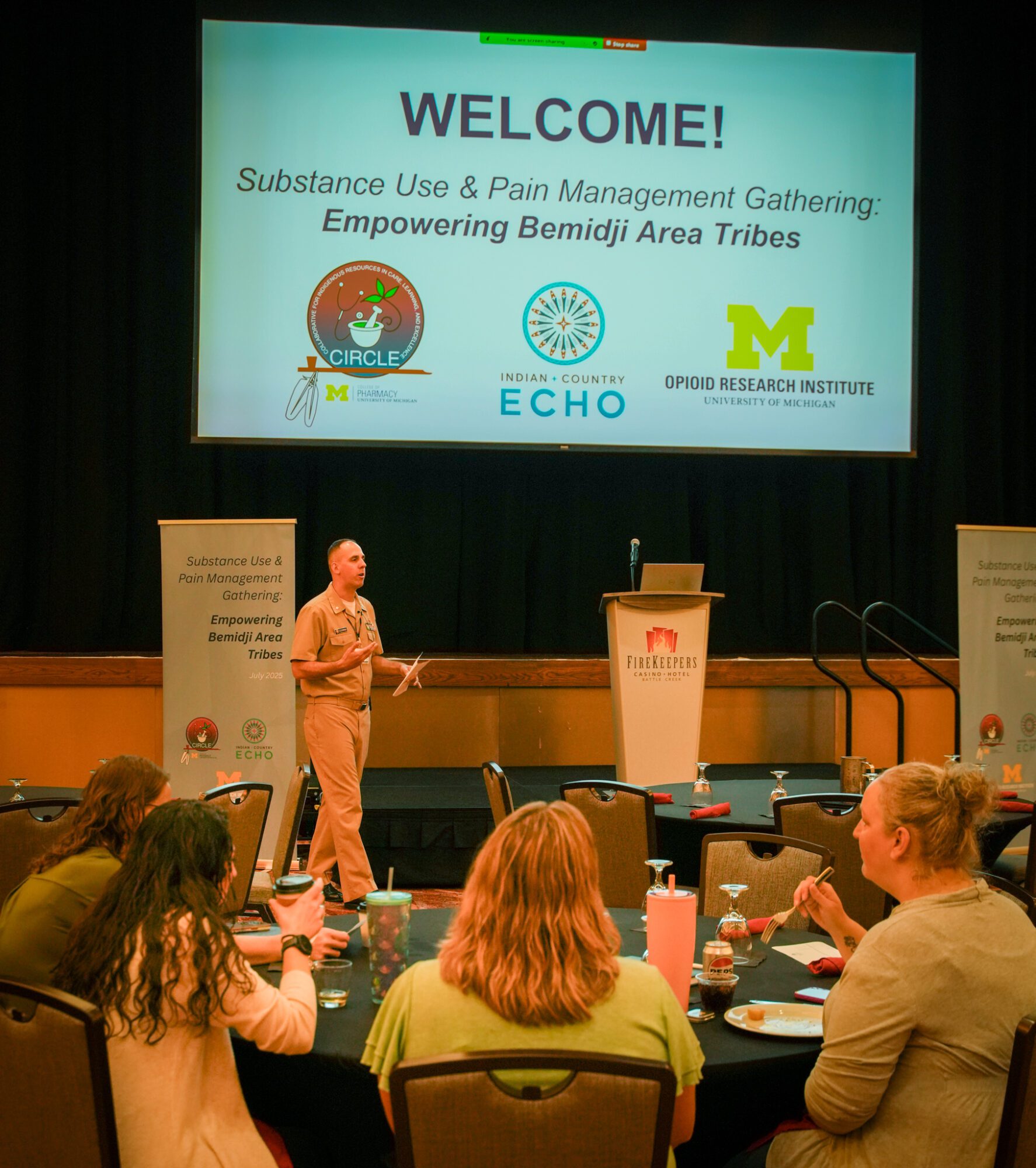 Conference room with attendees seated at round tables facing a stage, where a presenter stands near a podium beneath a large screen reading “WELCOME! Substance Use & Pain Management Gathering: Empowering Bemidji Area Tribes,” with CIRCLE, Indian + Country ECHO, and University of Michigan Opioid Research Institute logos.
