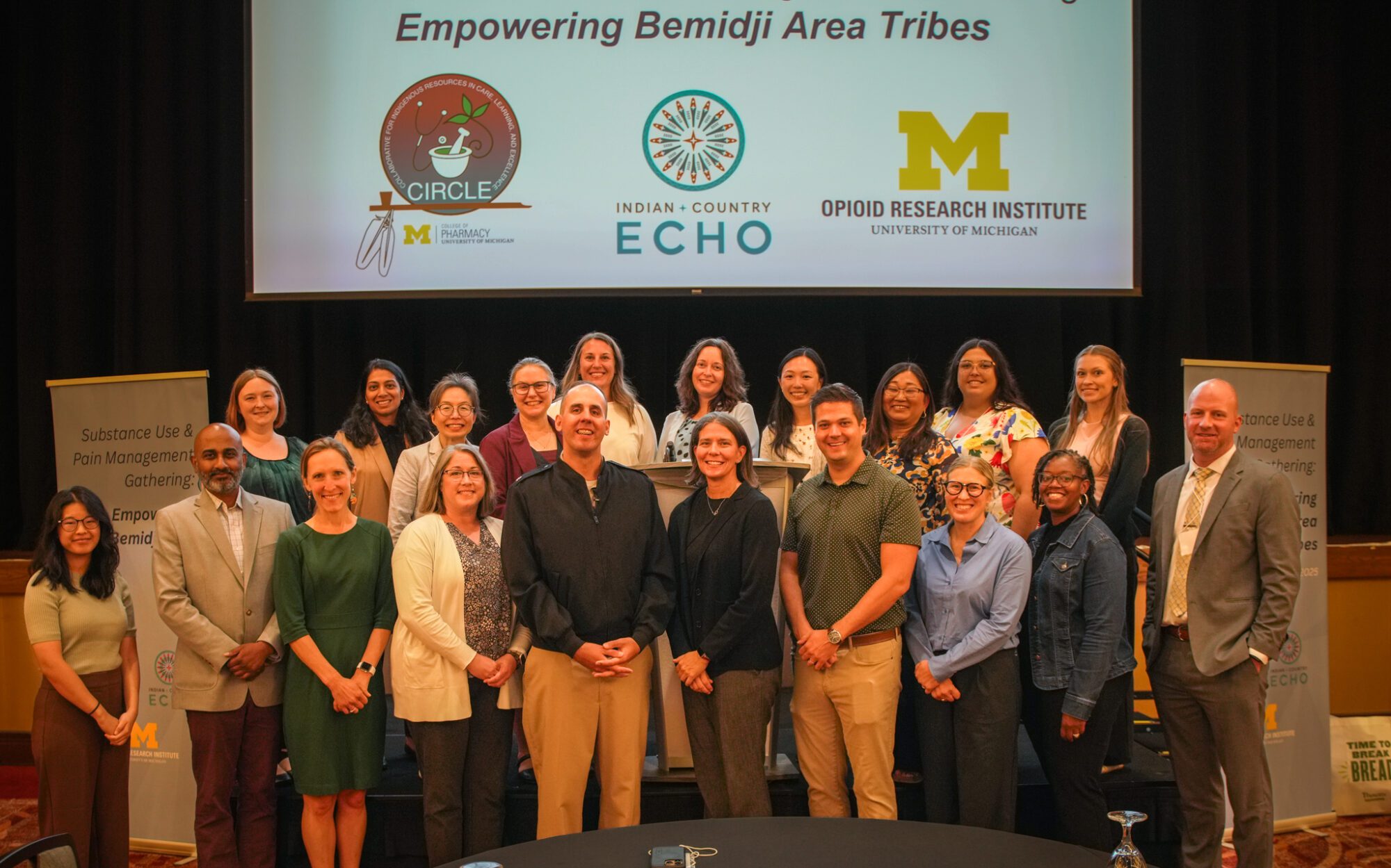 Group photo on a stage at a conference, with about 15–20 people standing in two rows beneath a banner reading “Empowering Bemidji Area Tribes” and logos for Circle, Indian + Country ECHO, and the University of Michigan Opioid Research Institute, flanked by podium signs about substance use and pain management.