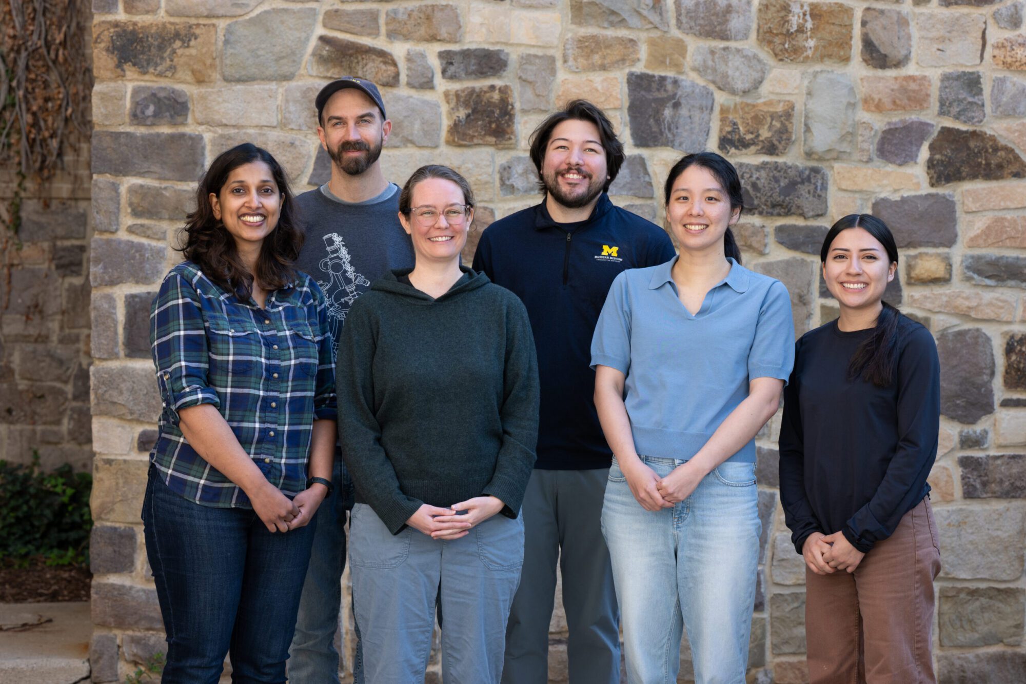 Group portrait of six people standing in two rows in front of a tan-and-gray stone wall; casual clothing, with one person wearing a navy University of Michigan pullover with a maize “M” logo.
