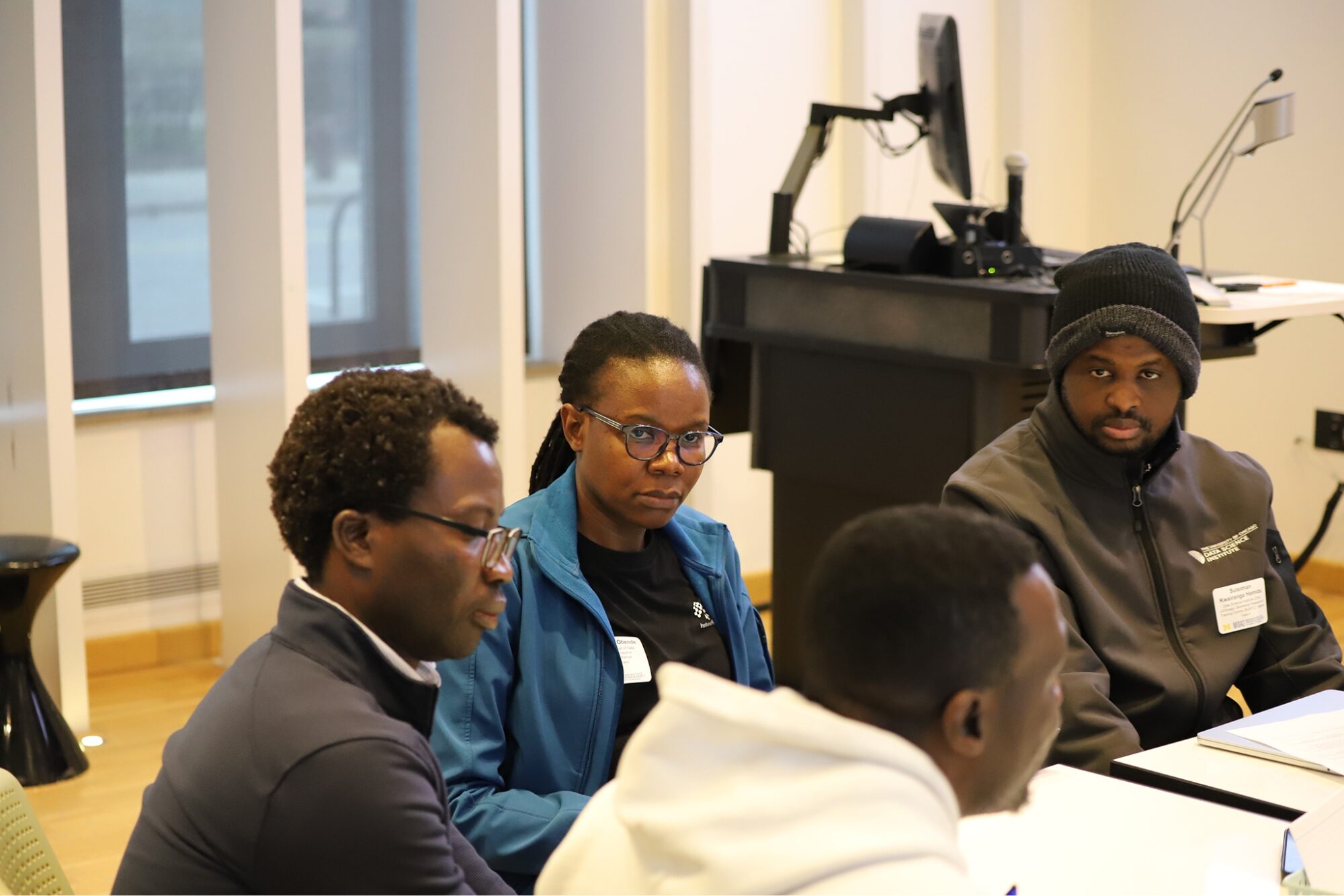 A small group of attendees sits at a table in discussion near a podium and presentation setup.