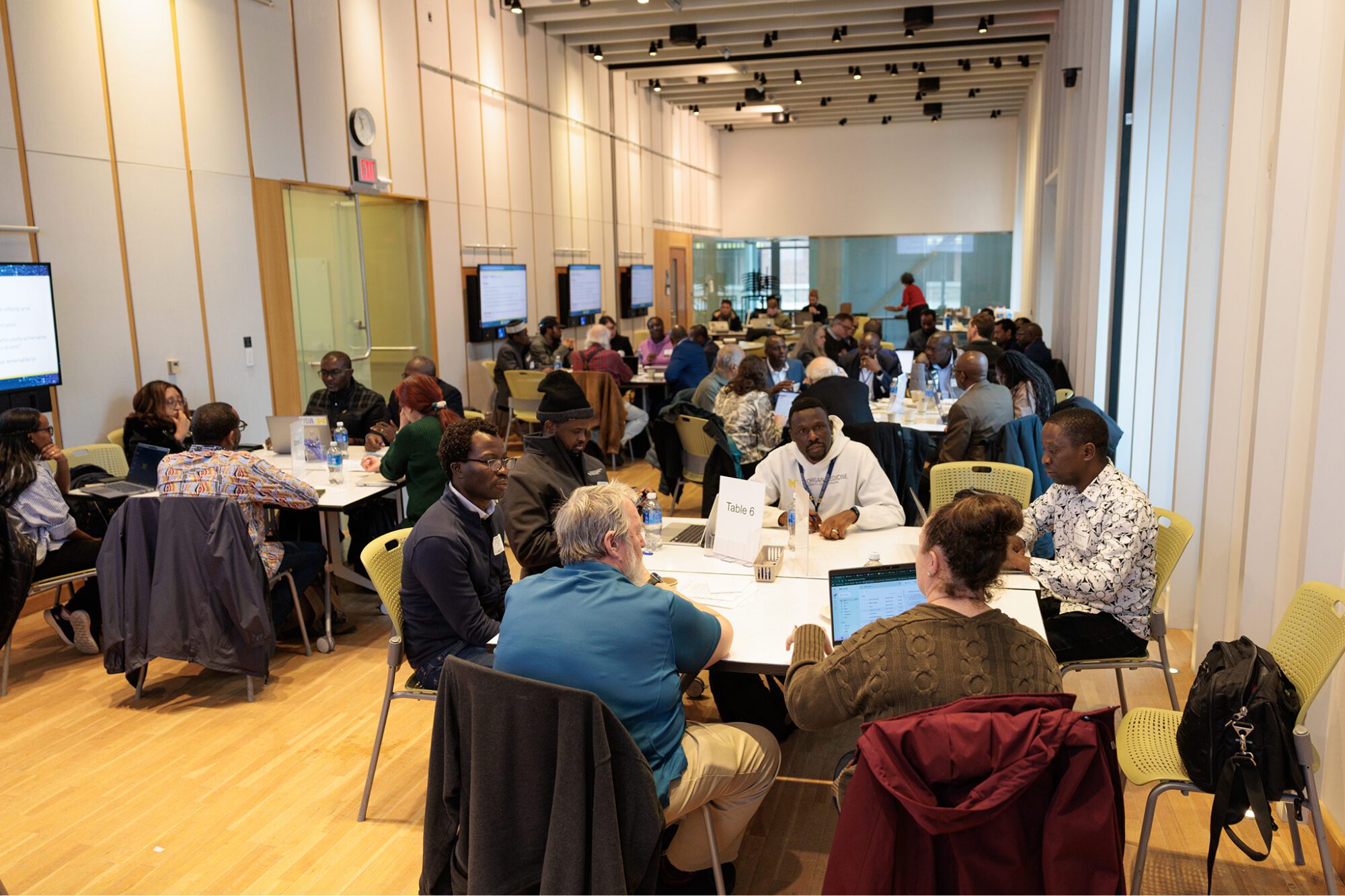 Wide view of a large workshop session with many people seated at round tables, talking and working; wall-mounted screens line the room.