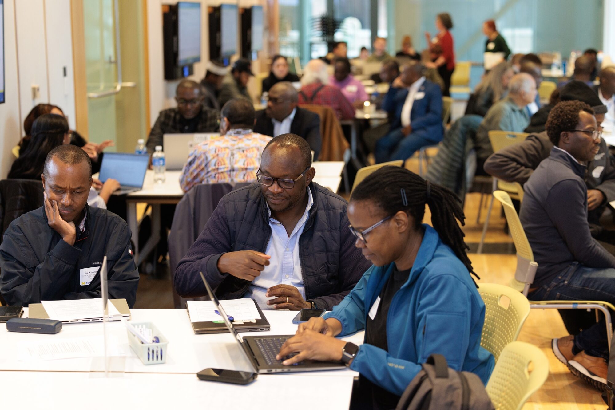  Participants sit at tables in a bright meeting room, working on laptops and discussing in small groups.