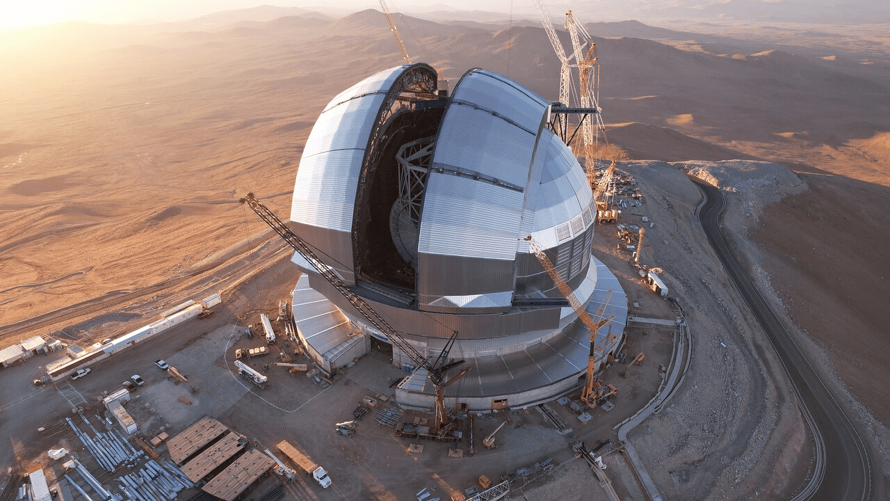 Aerial view of a large observatory dome under construction in a desert landscape, with segmented metal panels partially open and multiple cranes and construction vehicles surrounding the circular base near a winding road.