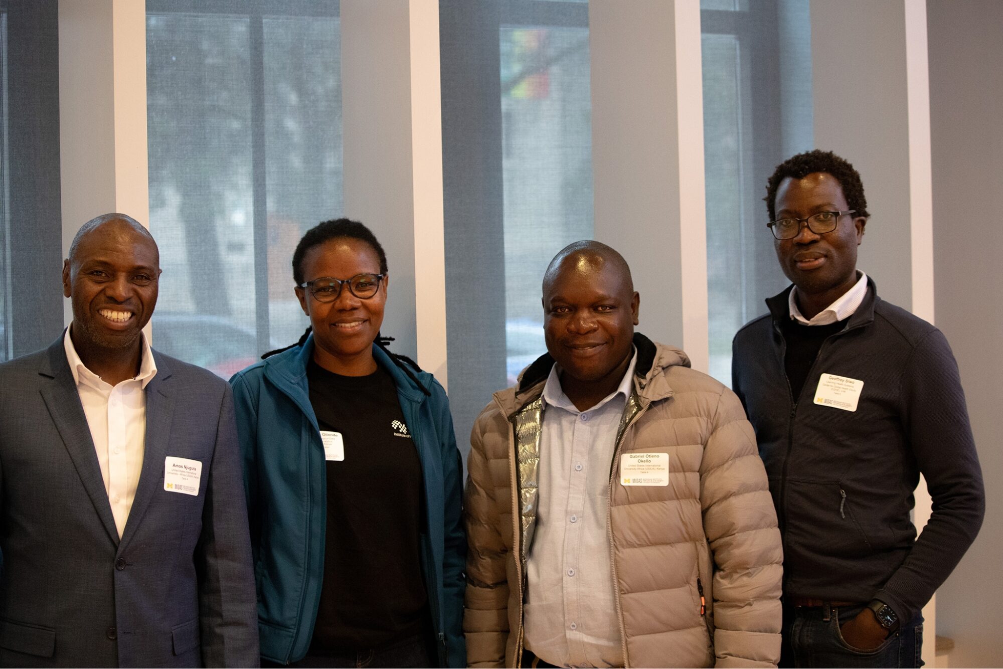 Four attendees stand side by side indoors for a group photo, wearing name tags and casual-to-business attire.