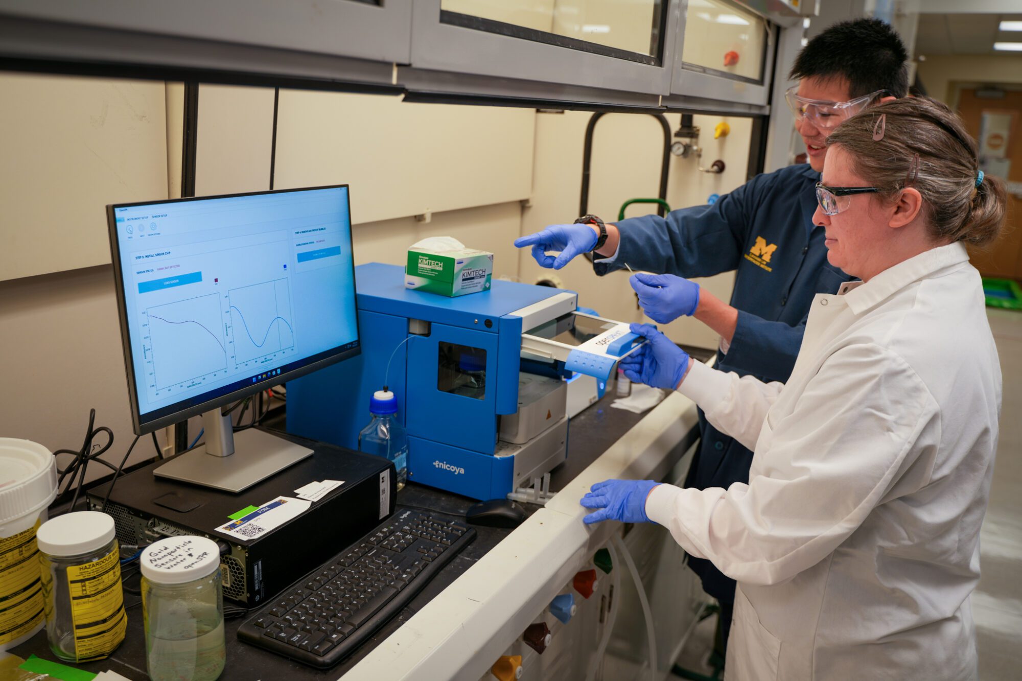 OVPR-Biointerfaces-Institute Two researchers in safety glasses and gloves operate laboratory equipment beside a computer displaying analysis graphs.