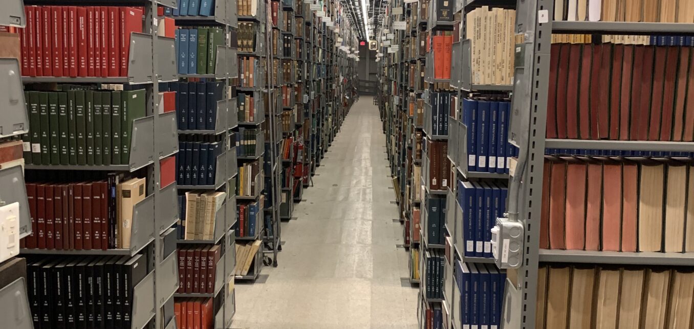 Rows of metal bookshelves filled with books and periodicals line both sides of a well-lit library aisle with a reflective tile floor.