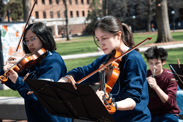 MCOMMONS-SpringSemester2026 Students perform violin and wind instruments outdoors beside music stands on campus.