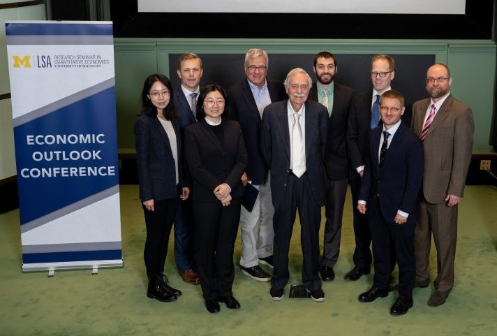 Ehrlich_Team Photo Nine conference participants pose beside a University of Michigan LSA banner reading “Economic Outlook Conference.”