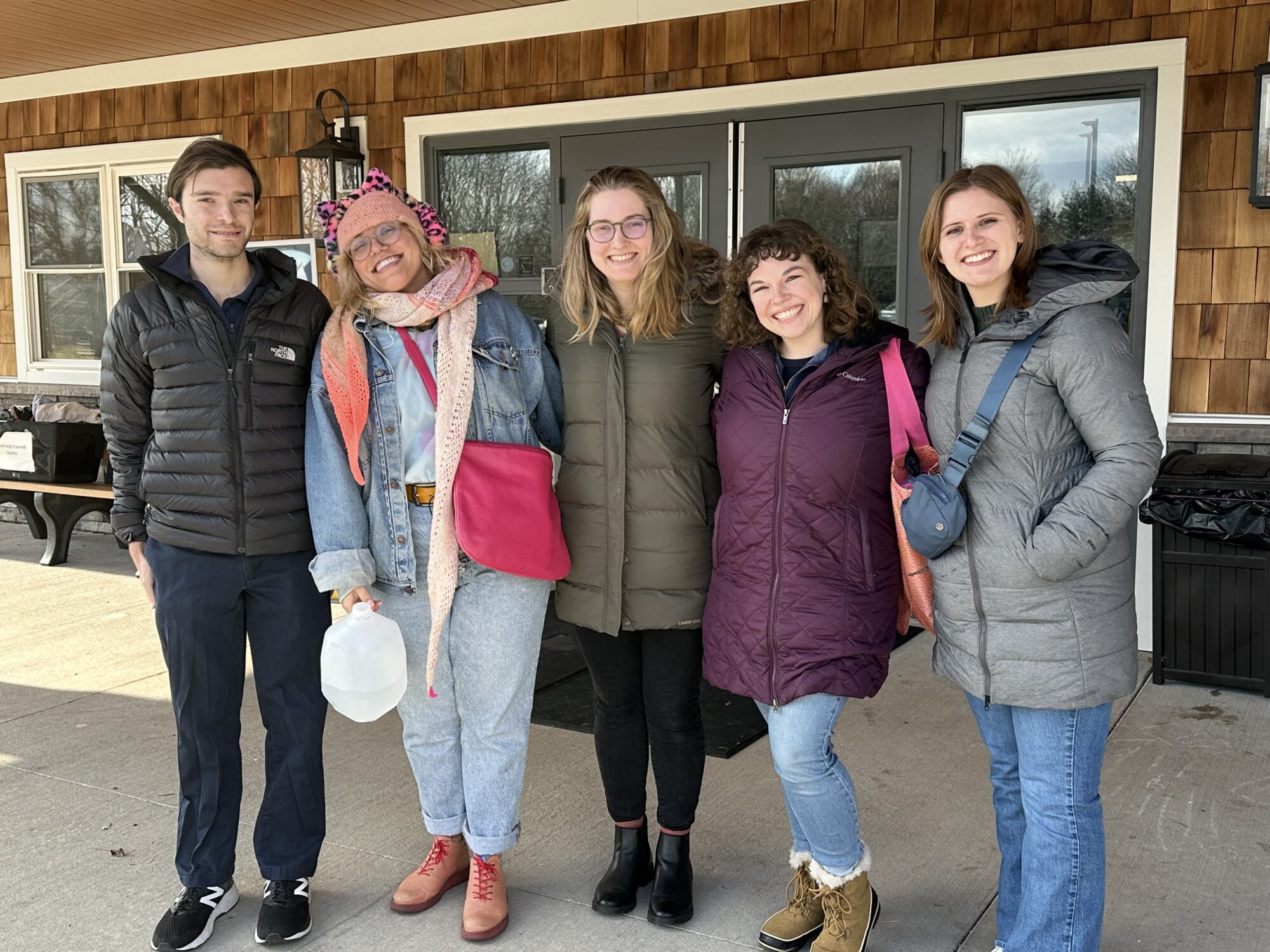 Five people in winter coats pose together outside a wood-sided building.