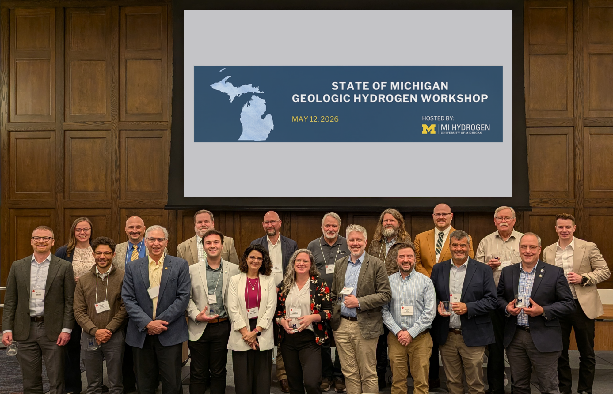 Nineteen attendees pose for a group photo beneath a screen reading “State of Michigan Geologic Hydrogen Workshop,” hosted by U-M Hydrogen on May 12, 2026. Several participants hold glass awards.