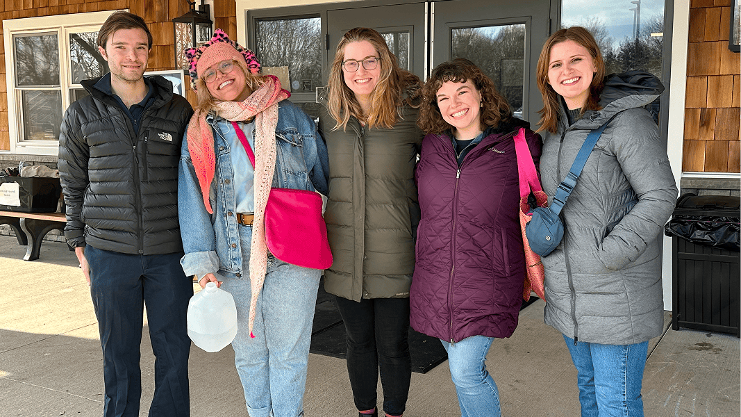 Five people in winter coats pose together outside a wood-sided building.
