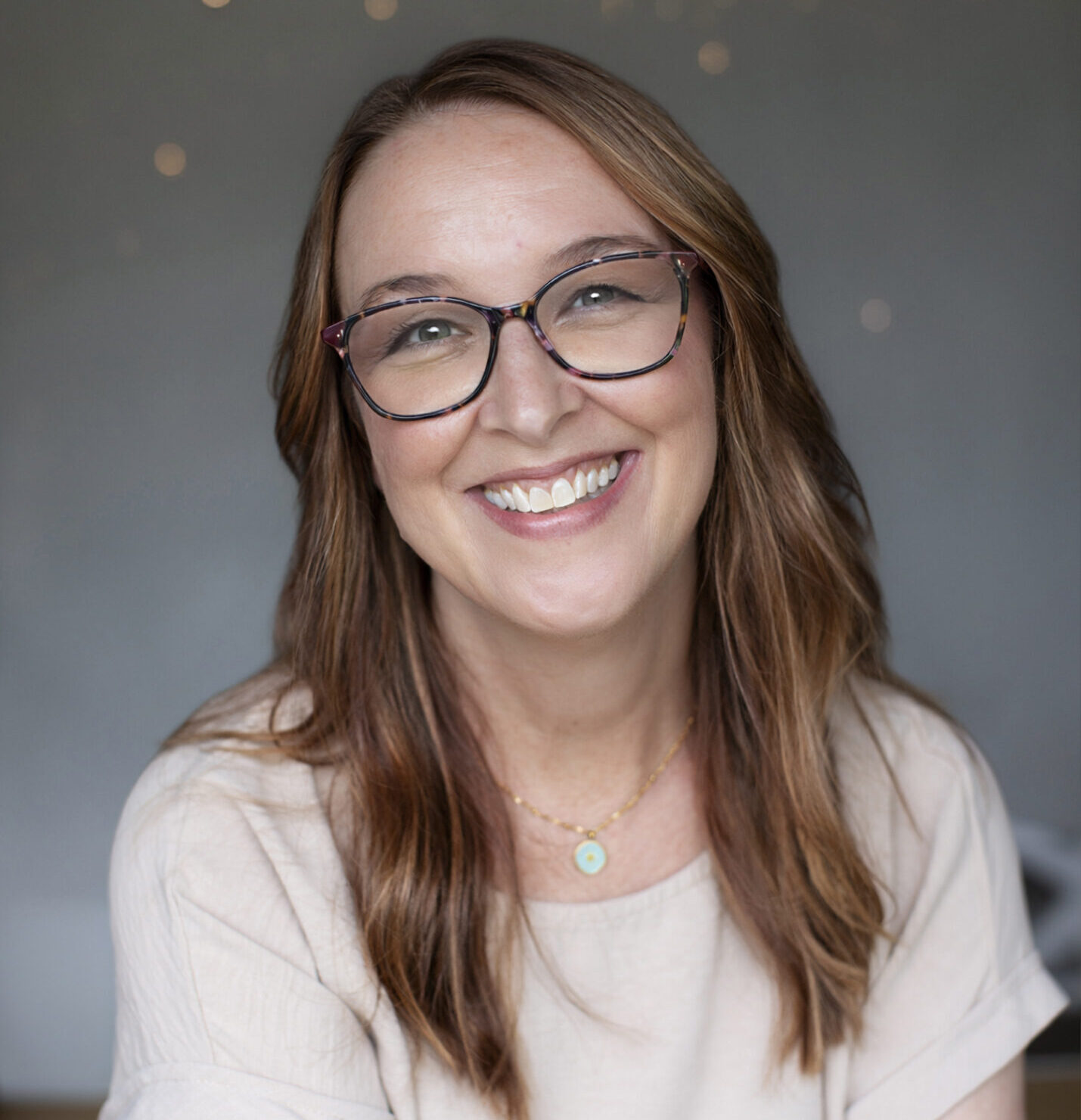 A smiling woman with long brown hair and patterned glasses poses in a beige top and pendant necklace against a softly blurred gray background with warm lights.